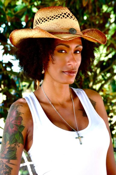 A woman with curly hair wearing a straw hat and a white tank top, displaying arm tattoos.